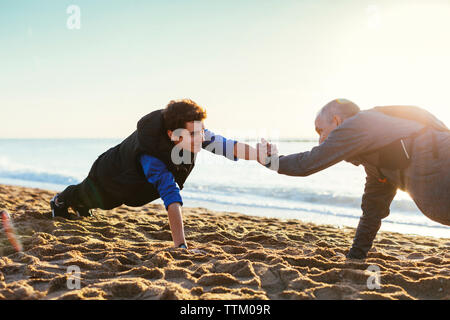 Vue latérale du père et fils faisant push-ups tout en maintenant les mains contre un ciel clair à la plage pendant le coucher du soleil Banque D'Images