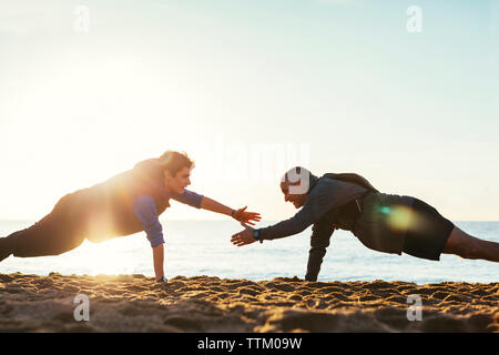 Vue latérale du père et fils faisant push-ups ensemble contre un ciel clair à la plage pendant le coucher du soleil Banque D'Images