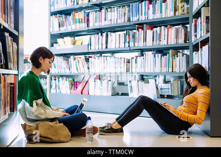 Amis de sexe féminin à travers l'étude d'ordinateurs portables tout en étant assis sur le plancher de la bibliothèque Banque D'Images
