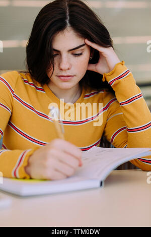 Woman writing on livre tout en sitting at table in library Banque D'Images