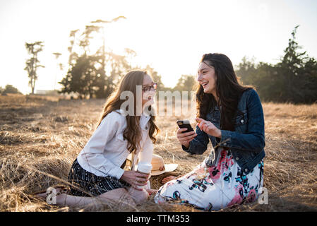 Heureux sisters en utilisant smart phone while sitting on grassy field against clear sky pendant le coucher du soleil Banque D'Images