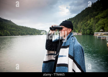 Vue de côté de l'homme regardant à travers des jumelles tout en se tenant par le lac contre ciel nuageux dans le parc national Olympic Banque D'Images