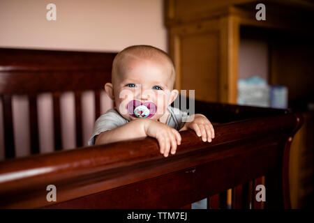 Portrait of happy baby girl sucking pacifier tout en se tenant dans un lit bébé Banque D'Images