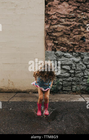 Playful girl jumping in puddle sur route contre mur Banque D'Images