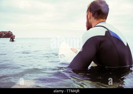 Vue arrière du surfeur hommes assis sur une planche de surf en mer contre ciel nuageux Banque D'Images