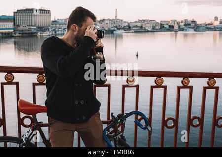 Photographier avec un appareil photo tout en cycliste avec permanent location sur pont sur la rivière en ville pendant le coucher du soleil Banque D'Images