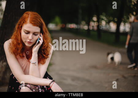 Jeune femme rousse talking on mobile phone while sitting in park Banque D'Images