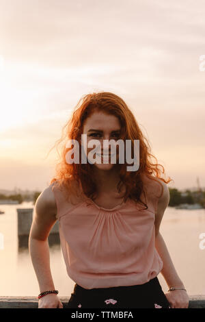 Portrait of smiling redhead woman leaning on railing at sunset Banque D'Images