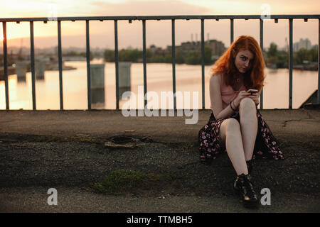 Jeune femme rousse à l'aide de smart phone while sitting on bridge en ville Banque D'Images