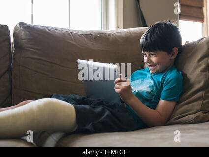 Smiling boy avec fracture de la jambe à l'aide de tablet computer while lying on sofa at home Banque D'Images