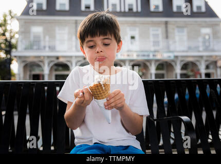 Cute boy eating ice cream cone tandis qu'assis sur un banc contre le bâtiment en ville Banque D'Images