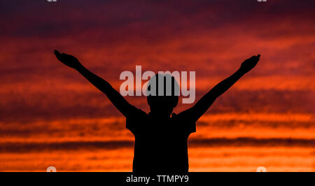 Silhouette garçon avec bras levés contre ciel dramatique pendant le coucher du soleil Banque D'Images