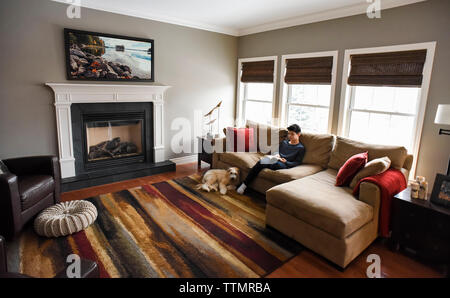 Boy reading sur canapé dans salon avec chien dans la journée. Banque D'Images