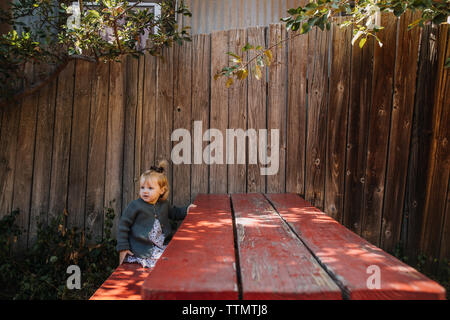 Jolie fille assise à une table de pique-nique rouge sous un pommier dans le soleil Banque D'Images