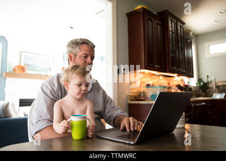 Travailler à la maison père assis à l'ordinateur dans la cuisine, tenant petit garçon Banque D'Images
