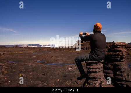 Vue arrière du male hiker photographing while sitting on rock against clear blue sky Banque D'Images