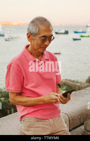 Senior man using smart phone alors que leaning on railing contre la mer Banque D'Images