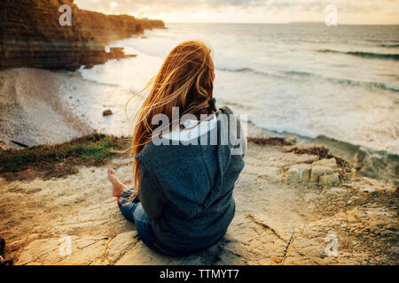 Vue arrière de la femme assis sur la falaise et à la vue pendant le coucher du soleil à Banque D'Images