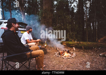 Amis assis sur les chaises de camping par feu de forêt Banque D'Images