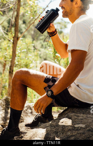 Vue latérale du male hiker eau potable lorsqu'assis sur un rocher dans la forêt Banque D'Images