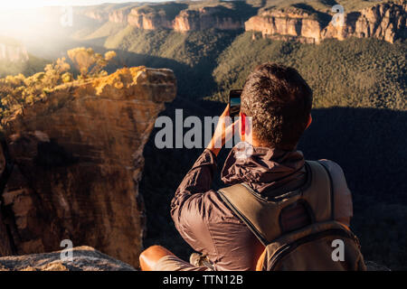 Vue arrière du sac à dos avec male hiker photographing tout en étant assis sur la montagne au cours de journée ensoleillée Banque D'Images