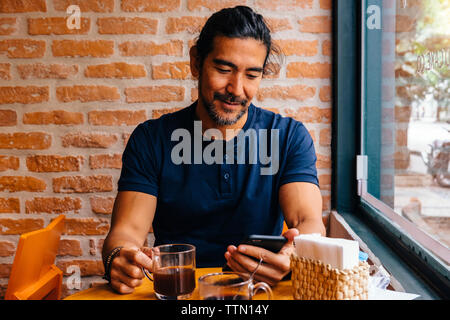 Homme mature avec du café noir à l'aide de smart phone against brick wall in cafe Banque D'Images