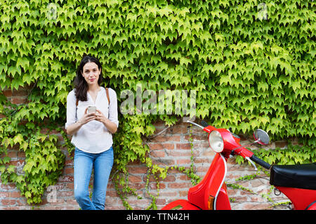 Portrait of woman holding smart phone en position debout par scooter contre mur de lierre Banque D'Images