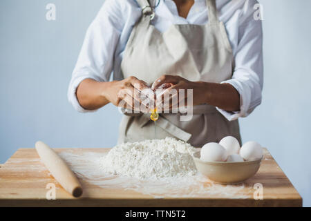 Midsection of woman breaking egg dans la farine sur la table en position debout contre le mur à la maison Banque D'Images