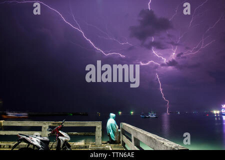 L'homme assis à pier par mer contre la foudre dans purple sky Banque D'Images