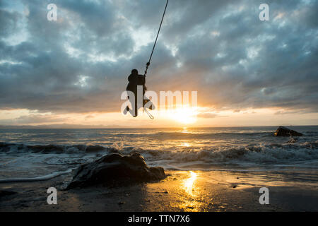 L'homme Silhouette accroché sur corde sur beach contre ciel nuageux pendant le coucher du soleil Banque D'Images