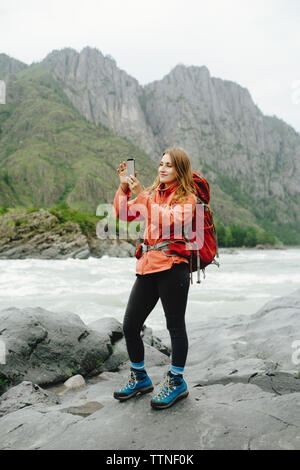 Female hiker photographing with mobile phone while standing on rocks at riverbank contre mountain Banque D'Images