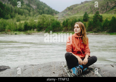 Thoughtful female hiker relaxing on rock at riverbank contre mountain Banque D'Images