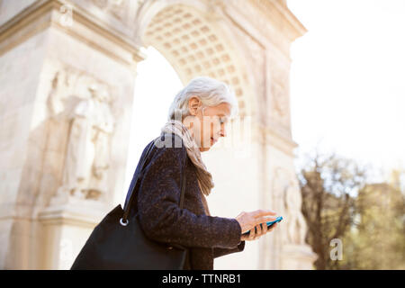 Vue latérale du senior woman using smart phone en position debout contre Triumphal Arch in city Banque D'Images