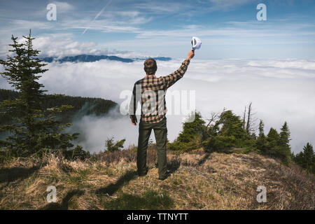 Vue arrière de l'homme tenant le chapeau et l'article sur la falaise contre cloudscape Banque D'Images