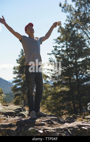 Happy man with arms outstretched holding Coffee cup en se tenant sur le rock contre le ciel en forêt Banque D'Images