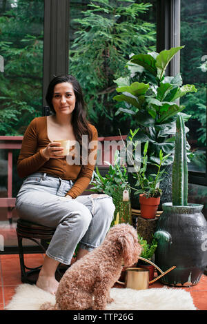 Portrait of young woman having drink while sitting par poodle et plantes en pot sur le porche Banque D'Images