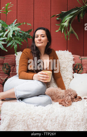 Portrait of young woman with poodle et verre reposant sur canapé par les plantes au porche Banque D'Images