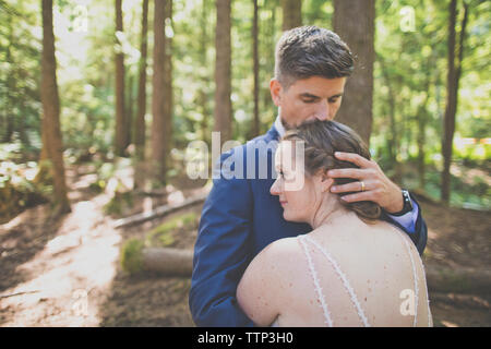 Romantic newlywed couple embracing while standing against trees in forest Banque D'Images