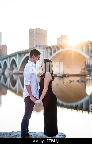 Heureux couple holding hands while standing par river contre ciel clair en ville Banque D'Images