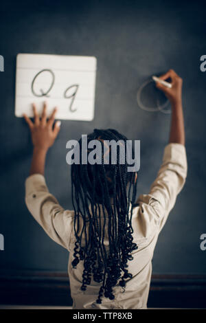 Vue arrière d'écolière avec cheveux tressés en alphabet écrit Q on blackboard Banque D'Images