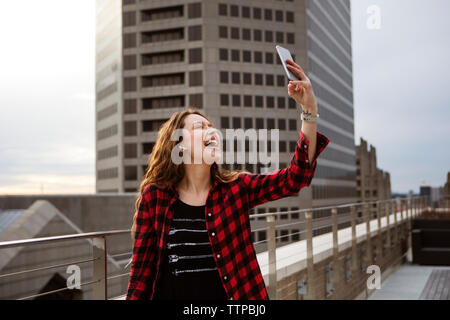 Jeune femme gaie en tenant debout sur terrasse selfies contre le bâtiment Banque D'Images