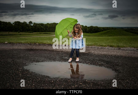 Girl holding umbrella en marchant en flaque sur route contre des nuages orageux Banque D'Images