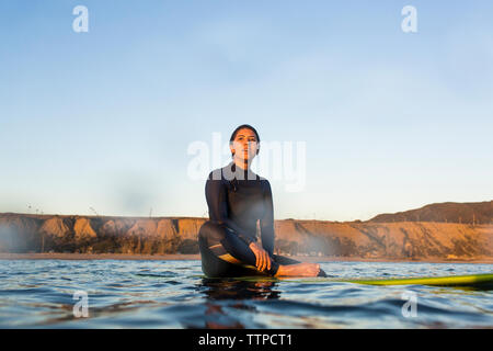 Jeune femme assise sur une planche de surf en mer contre ciel clair Banque D'Images