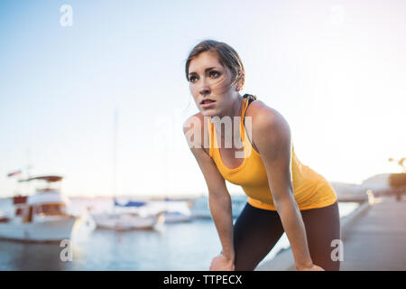 Athlète féminine fatigué debout avec les mains sur les genoux par harbour contre ciel bleu clair Banque D'Images