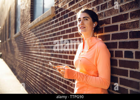 Thoughtful woman sportive tout en écoutant de la musique via smart phone par brick wall Banque D'Images