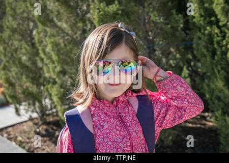 Close-up portrait of girl avec sac à dos porter des lunettes de soleil en position debout contre des plantes au cours de journée ensoleillée Banque D'Images