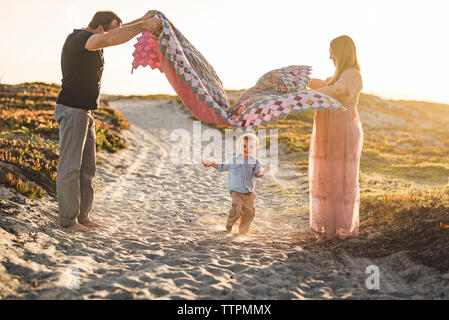 Vue latérale des parents holding couverture sur mignon fils d'exécution sur la plage de sable à contre ciel clair pendant le coucher du soleil Banque D'Images