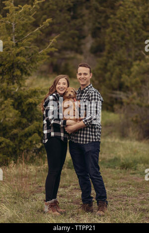 Portrait of smiling young couple carrying dog en se tenant sur le terrain herbeux Banque D'Images