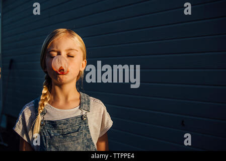 Fille aux yeux fermés soufflant bubble gum en position debout par wall Banque D'Images