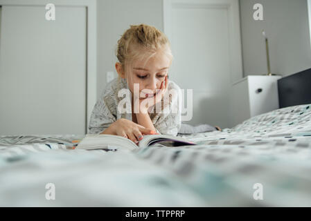 Girl with hand on chin reading book while lying on bed at home Banque D'Images